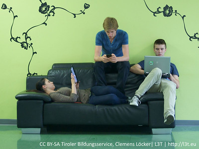 Three students on a sofa using different devices. The first student is looking at a tablet. The second student is on their mobile phone. The third student is on their laptop.